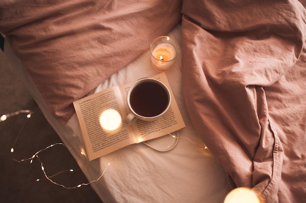 Kopje verse warme koffie die op een open papieren boek blijft met brandende kaars en gloeiende kerstverlichting in bed close-up. Winter vakantie seizoen. Goedemorgen. Selectieve aandacht.