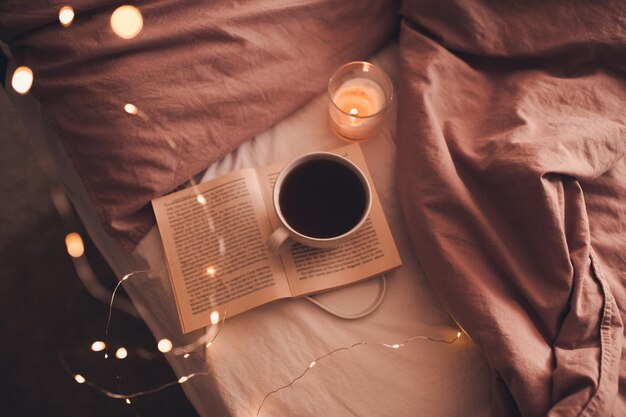 Kopje verse warme koffie die op een open papieren boek blijft met brandende kaars en gloeiende kerstverlichting in bed close-up. Winter vakantie seizoen. Goedemorgen. Selectieve aandacht.