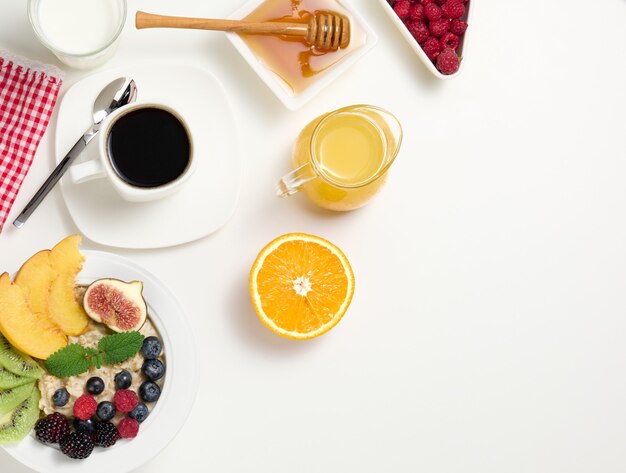 Kopje zwarte koffie, een bord havermout en fruit, honing en een glas melk op een witte tafel, een gezond ochtendontbijt