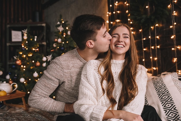 Foto koppel op het bed in de slaapkamer. donker interieur. nieuwjaar en kerstmis. knuffels en kussen. dol zijn op. witte trui en hoge sokken. romantische ontmoeting. liefhebbers van een date.