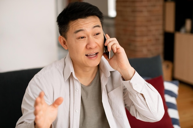 Koreaanse man van middelbare leeftijd zittend op de bank thuis met telefoongesprek en gebaren met de hand