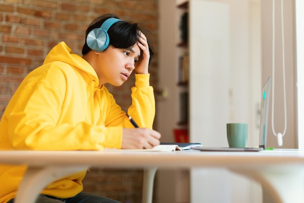 Koreaanse tienerjongen op laptop die online leert met oortelefoons binnenshuis