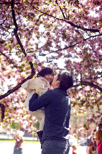 父を楽しんでいる韓国の家族は、咲く桜の庭で彼の腕の中で娘を持ち上げます