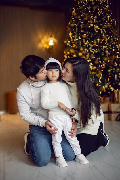 한국 가정의 엄마 아빠와 딸 아이가 집에 있는 스튜디오에서 화환을 들고 크리스마스 트리에 앉아 있다