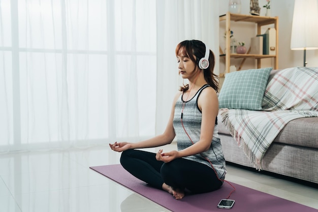 Korean female in sportswear is listening to peaceful music from headphones with her smartphone while meditating on the yoga mat at a bright home interior.