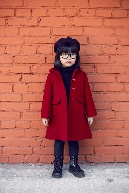 Korean girl in a red coat and cap and round glasses stands on\
red brick wall the street in autumn