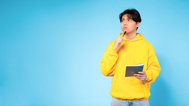 Korean guy with pen and textbook thinking on blue background