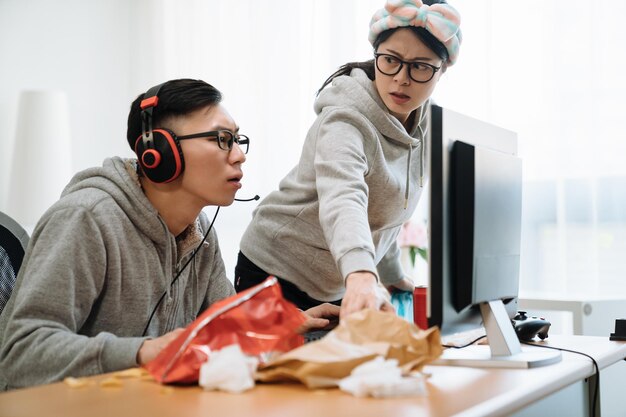 korean man in headphones playing online game on computer while angry wife standing and staring at him. Woman doing chores while man resting in messy home on weekends. female clean up trash on desk