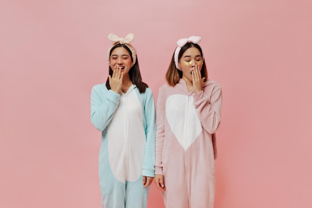 Kortharige Aziatische meisjes in hoofdbanden en cosmetische ooglapjes gapen Cure brunette vrouwen in pyjama poseren op roze achtergrond