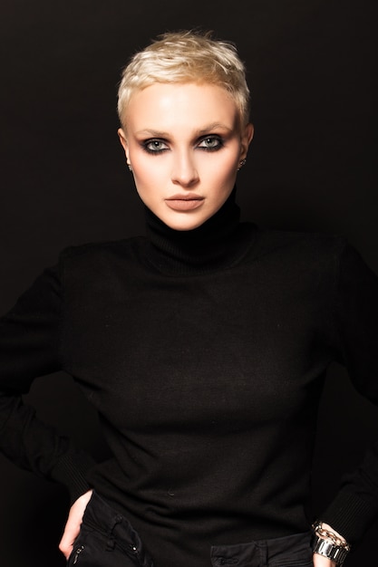 Kortharige blonde in zwarte kleding op een zwarte muur.