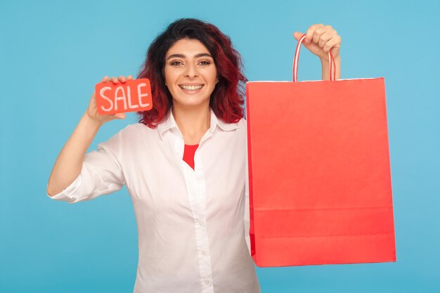 Korting in modewinkel Leuke vrolijke hipster vrouw met mooie rode haren met verkoop inscriptie en boodschappentassen genieten van zwarte vrijdag lage prijzen binnen studio opname geïsoleerd op blauwe achtergrond