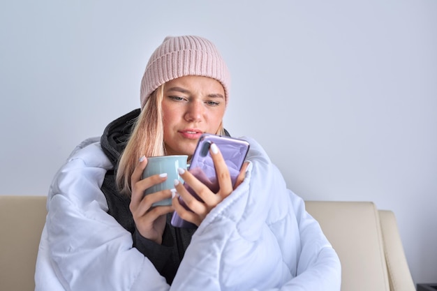 Koud herfst winterseizoen, tienermeisje in wollen gebreide muts zittend op de bank onder warme deken opwarmen met mok hete thee en smartphone gebruiken