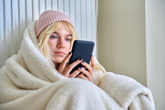 Koud seizoen bevroren tiener in hoed onder deken zittend in de buurt van een verwarmingsradiator met smartphone