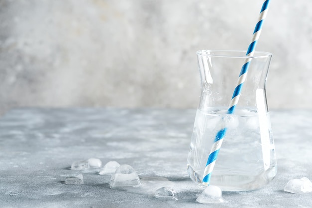 Koud verfrissend natuurlijk water in een glaskruik met plastic stro met ijsblokjes op een marmeren grijze achtergrond.