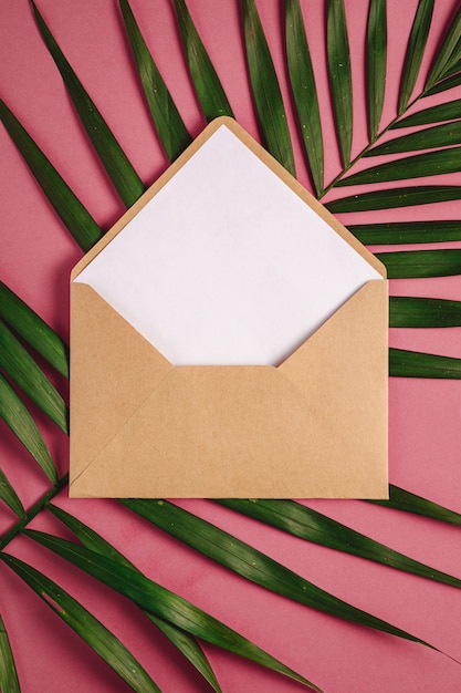 Kraft brown paper envelope with white empty card on palm leaves, pink red background, mockup blank letter