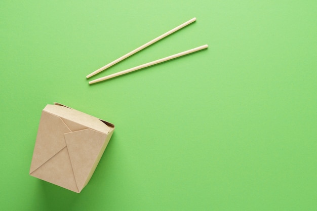 Kraft paper or cardboard container and chopsticks on green background. Food delivery concept. Copy space.