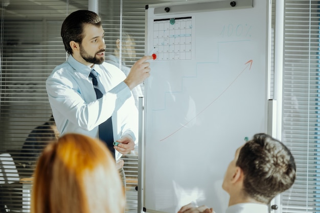 Krappe deadlines. Knappe jonge mannelijke baas permanent in de buurt van het whiteboard en wijzend op de kalender die erop is vastgemaakt terwijl hij praat over de deadlines van het project