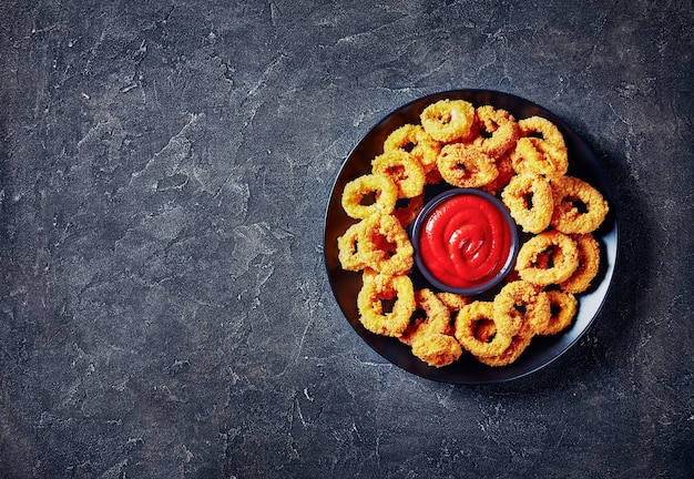 Krokante calamaresringen, gefrituurde gepaneerde inktvisringen geserveerd met tomatensaus op een zwarte plaat op een betonnen tafel, horizontaal zicht van bovenaf, vrije ruimte, flatlay