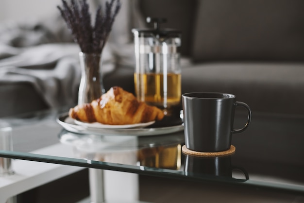 Kruidenthee in een zwarte mok en croissant geserveerd op een glazen salontafel in de woonkamer