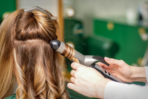 Krullen maken met krultangen. kapper maakt een kapsel voor een jonge vrouw met lang rood haar in een schoonheidssalon.