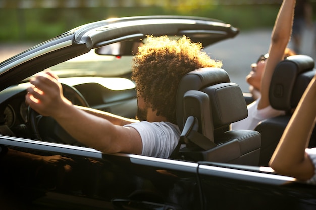 Krullend donkerharige jongeman zit aan het stuur van een zwarte cabriolet en zijn vrienden zitten naast hem op een warme zonnige dag. .