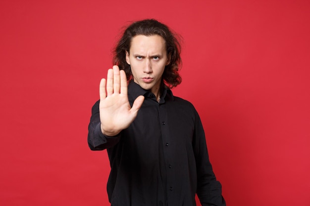 Krullend langharige man in zwart shirt poseren geïsoleerd op rode muur achtergrond studio portret. Mensen oprechte emoties levensstijl concept. Mock-up kopieerruimte Kijkende camera met stopgebaar met palm