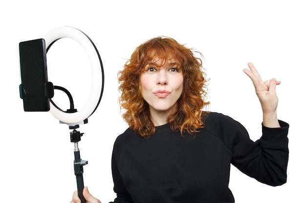 Krullend roodharig meisje dat naar de camera kijkt met een kussende pose en een lichte ring vasthoudt met een mobiele telefoon op een witte achtergrond