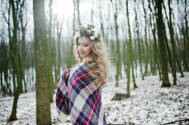 Krullend schattig blondemeisje met kroon in geruite plaid bij sneeuwbos in de winterdag.