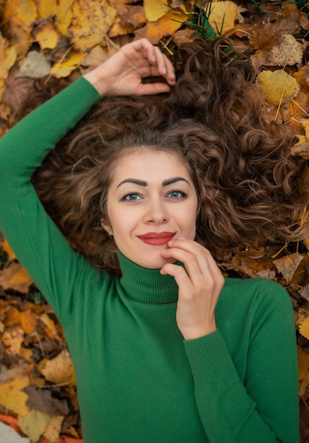 Krullend schattig meisje met open ogen liggen in de herfstbladeren, hand in de buurt van gezicht en glimlach