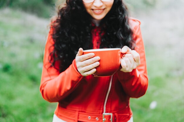 Krullende brunette vrouw, gekleed in een rood leren jack en met een rode mok met warme koffie in de natuur.