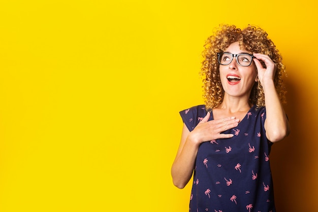 Krullende jonge vrouw kijkt verbaasd op terwijl ze een bril vasthoudt op een gele achtergrond