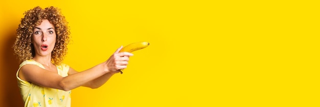 Krullende jonge vrouw met een banaan als een pistool op een gele achtergrond Banner