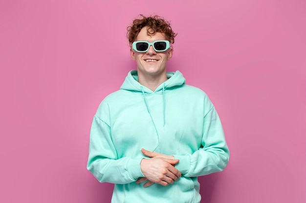 krullende man in mint hoodie en zonnebril glimlacht op roze geïsoleerde achtergrond man in blauwe sweatshirt