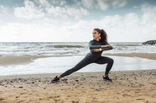 Krullende sportvrouw in sportkleding die lichaamsopwarming doet of zich uitstrekt op het strand Gezonde levensstijl