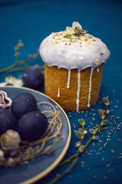 Kulich with sugar glaze decoration design blue easter eggs lie on a plate with a willow on fabric background Paska Easter Bread