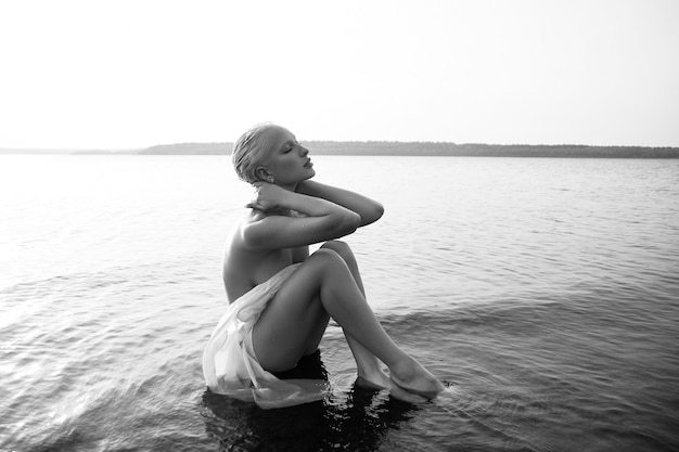 Kunst naakt sexy blonde met kort kapsel zit in het water op het strand van het meer bij zonsondergang. Nat haar en een vrouwenlichaam. Afgelegen strandvakantie. Zwart en wit