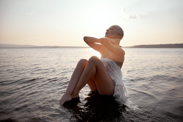 Kunst naakt sexy blonde met kort kapsel zit in het water op het strand van het meer bij zonsondergang. Nat haar en een vrouwenlichaam. Afgelegen strandvakantie