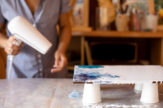 Kunstenaar maakt foto op tafel in haar atelier. Ze mengt kleuren door ze met een föhn te föhnen. Binnen schilderen. Creativiteit en vormgeving. Hobby's en ambachten. Vrijheid en creativiteit. Levensstijl.