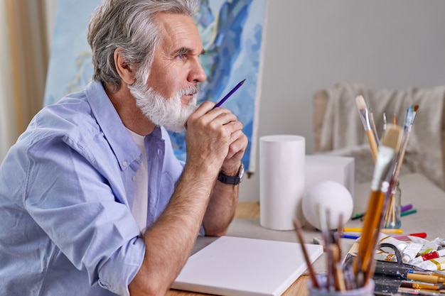 Kunstenaar zit achter tafel en denkt wat te tekenen, schilderen. oudere mannen gebruiken potlood voor tekeningen