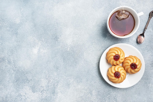 Kurabye shortbread cookies with berry jam in a plate and a cup of tea. Delicious dessert, gray concrete background with copy space.
