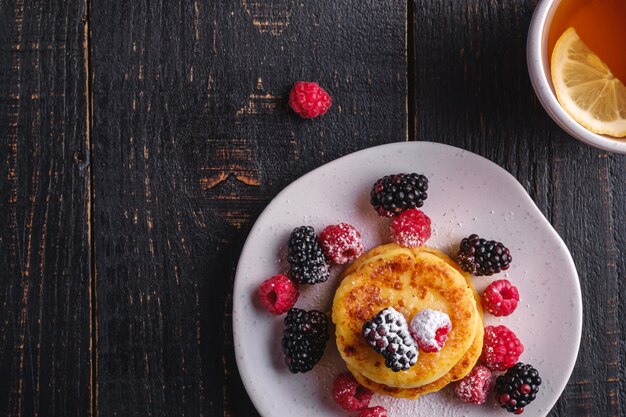 Kwark pannenkoeken, kwark beignets dessert met frambozen en bramen bessen in plaat in de buurt van hete thee beker met schijfje citroen op donkere zwarte houten tafel, bovenaanzicht kopie ruimte