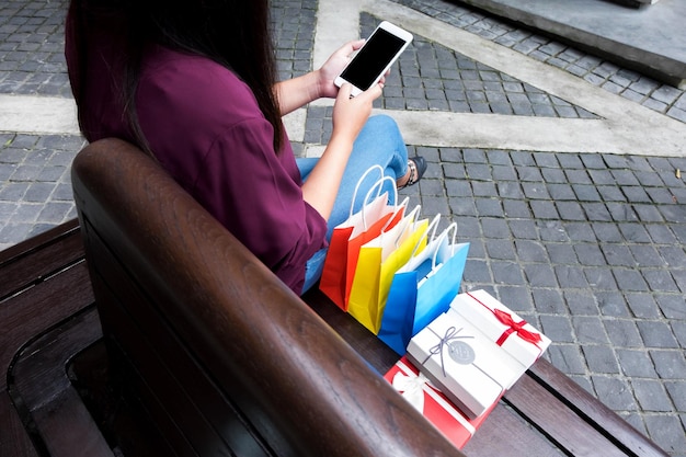 Foto laag gedeelte van een jonge vrouw met winkeltassen die een mobiele telefoon gebruikt terwijl ze op een bank zit