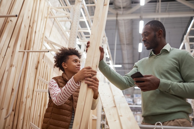 Laag hoekportret van Afro-Amerikaanse vader en zoon die samen winkelen in de ijzerhandel, focus op man die houten planken kiest voor constructie of woningverbetering