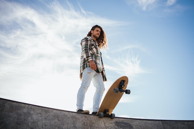 Laag hoekportret van een langharige coole man met zijn skateboard op de rand van het zwembad