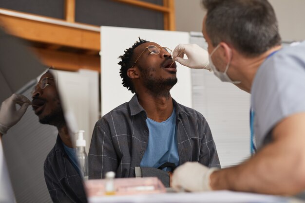 Laag hoekportret van jonge Afro-Amerikaanse man die covid-test doet in vaccinatiecentrum of kliniek, kopieer ruimte