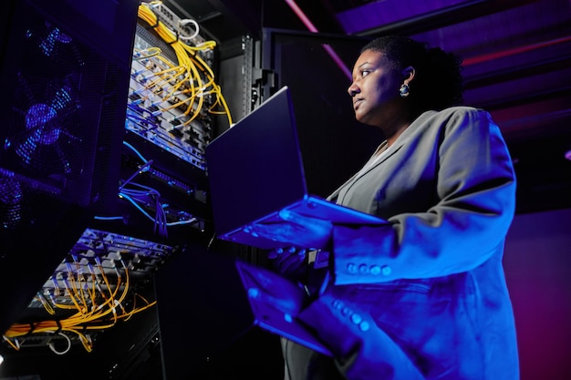 Laag hoekportret van vrouwelijke netwerkingenieur die servers opzet in datacenter verlicht door neon