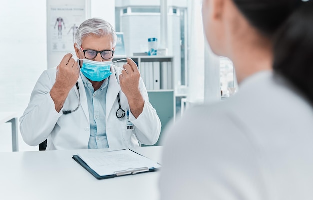 Laat me dit eerst doen Shot van een volwassen arts die een gezichtsmasker opzet tijdens een consult met zijn patiënt in de kliniek