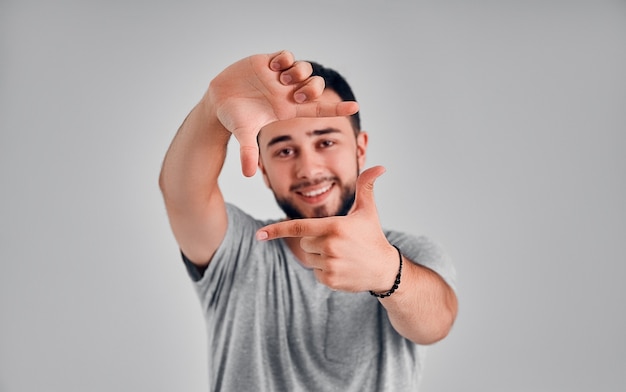 Laat me je foto maken. Portret van aantrekkelijke emotionele witte mannelijke vinger inlijsten en glimlachen naar de camera, staande tegen een grijze achtergrond, proberen een goede scène te creëren tijdens het maken van een film