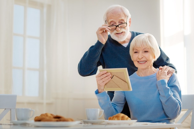 Laat ons herdenken. Positief opgetogen gepensioneerde die een glimlach op zijn gezicht en een rimpelend voorhoofd houdt terwijl hij zijn vrouw omhelst