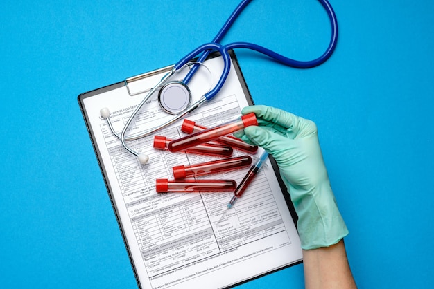 실험실 기술자 조수 또는 의사가 빈 양식 클립 보드 위에 혈액 검사 튜브를 들고 고무 또는 라텍스 장갑을 끼고 있습니다.